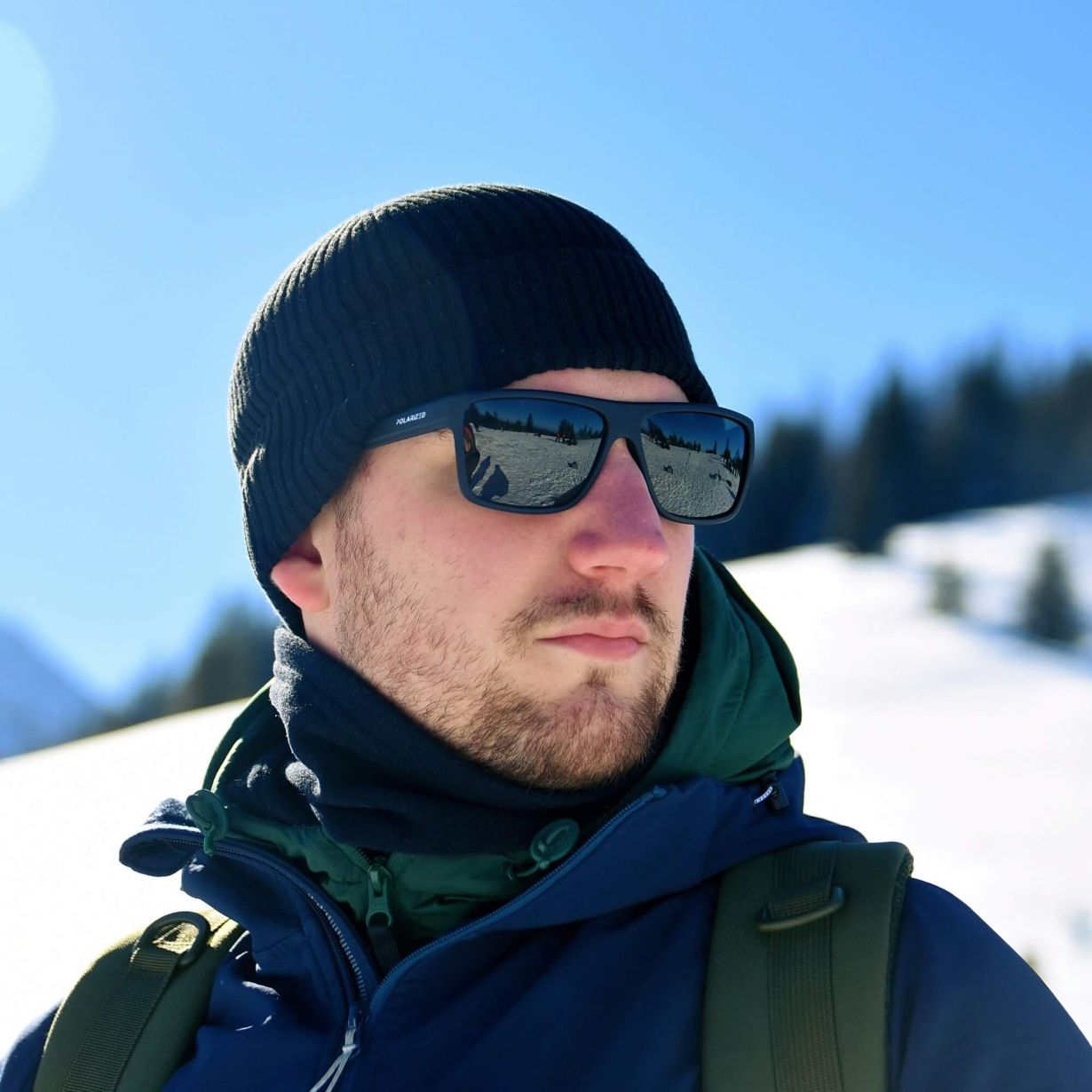 Uma foto de um homem com óculos de sol e uma touca azul, usando um casaco de neve também azul, em um fundo de montanhas e céu azul.